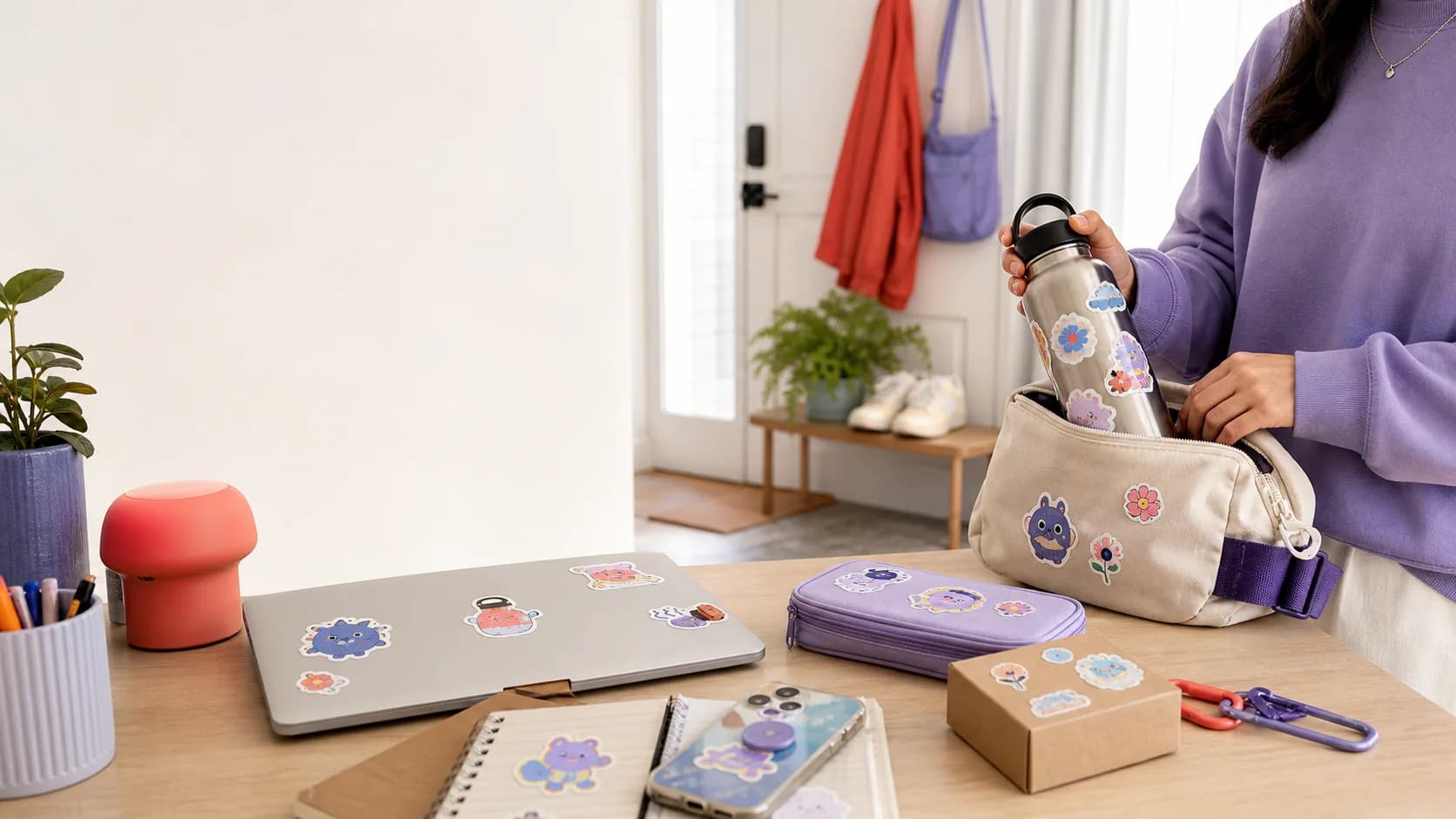 A person packing a sticker-covered bottle beside a laptop, phone, notebook, travel case, and package
